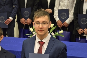 Paweł Dziwoki - uczestnik Olimpiady Geograficznej w garniturze trzyma dyplom na tle grupy innych finalistów. Wszyscy ubrani formalnie, w tle stół z niebieskim obrusem i kwiatami. Widoczne logo „Olimpiada Geograficzna”.
