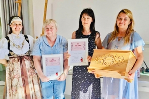 Cztery kobiety pozują w jasnym wnętrzu. Po lewej osoba w bogatym stroju ludowym, obok dwie laureatki trzymają dyplomy z napisami „III MIEJSCE” i „II MIEJSCE”. Po prawej kobieta w błękitnej sukience prezentuje duży, rzeźbiony drewniany kufer z datą 2025. Atmosfera uroczysta i radosna.