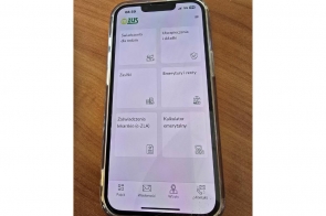 Smartfon z włączoną aplikacją mZUS pokazuje czytelne kafelki z kategoriami takimi jak świadczenia, zasiłki czy emerytury. Leży na drewnianym blacie, w przezroczystym etui, a ekran jest dobrze oświetlony i wyraźny.