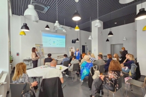 W nowoczesnej sali warsztatowej prowadząca prezentuje slajd przy ekranie.Uczestnicy siedzą przy stołach i na krzesłach, słuchają i notują; na stołach widać markery i materiały. W tle druga prowadząca w niebieskim żakiecie.