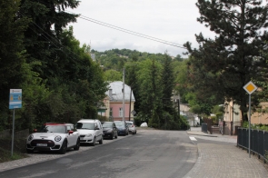 Wąska ulica otoczona zielenią, po lewej szereg zaparkowanych aut, w tym biały Mini z czerwonym dachem. Po prawej ogrodzenia i znak pierwszeństwa, w tle pastelowe domy oraz zalesione wzgórza pod pochmurnym niebem.