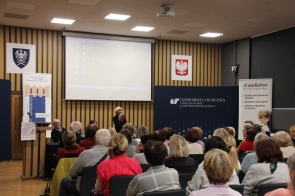 W sali konferencyjnej siedzi liczna grupa osób zwróconych w stronę mównicy i panelu prelegentów. Na scenie stoi kobieta mówiąca do mikrofonu, a za nią widać duży ekran projekcyjny oraz banery informacyjne. Pomieszczenie ma drewniane, pionowe panele na ścianach i emblematy państwowe oraz uczelniane.