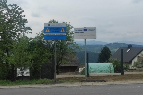 Dwa znaki informacyjne ustawione przy drodze. Jeden z nich informuje o prowadzonych pracach budowlanych i jest oznaczony nazwą Powiat Cieszyński, drugi zawiera logo Interreg Polska-Słowacja oraz informację o współfinansowaniu przez Unię Europejską. W tle widoczne są drzewa, zabudowania oraz pagórkowaty krajobraz.