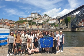Grupa młodzieży i kilka dorosłych osób pozujących nad rzeką w Portugalii. W tle widać kolorowe kamienice i most. Jedna z dziewczyn trzyma flagę Unii Europejskiej. Zdjęcie zrobiono w słoneczny dzień.