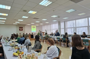 Na zdjęciu widać salę konferencyjną, w której przy długich stołach siedzą uczestnicy spotkania. Na stołach ustawione są przekąski i napoje. Z przodu sali jedna osoba przemawia przy mównicy, a obok niej stoi prowadzący. W tle widoczny jest ekran z prezentacją oraz godło Polski.
