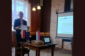 Kobieta stojąca przy stole trzyma dokument. Na stole znajduje się projektor, laptop oraz małe flagi Polski, Unii Europejskiej i Słowacji. Obok ustawiony jest ekran z wyświetloną prezentacją programu Interreg Polska-Słowacja. W tle widoczne są krzesła, okno z zasłonami i oświetlenie sufitowe.