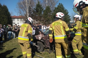 Strażacy z Ochotniczej Straży Pożarnej w pełnym umundurowaniu przeprowadzają pokaz ratownictwa technicznego, używając nożyc hydraulicznych przy uszkodzonym samochodzie. W tle stoi grupa uczniów obserwujących działania.