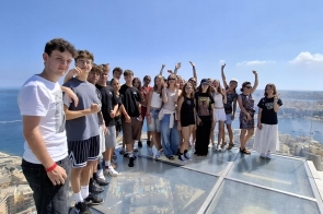 Grupa nastolatków i dorosłych stoi na szklanym tarasie widokowym wysoko nad nadmorskim miastem. Słoneczny dzień, błękitne niebo i rozległe morze z zatoką oraz łódkami w dole. Wszyscy uśmiechnięci, część macha do aparatu, luźne, letnie  stroje.