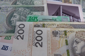 Polskie banknoty o nominałach 100 i 200 złotych leżą obok zielonej legitymacji emeryta z logo ZUS oraz kilku dokumentów urzędowych. W tle widoczne są urzędowe pisma i zdjęcie budynku.