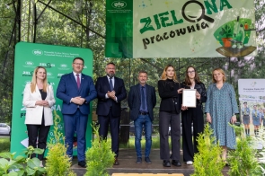 Grupa uśmiechniętych osób stoi na scenie pod banerem „Zielona Pracownia”. Wśród nich są przedstawiciele instytucji i szkół, jedna z kobiet trzyma dyplom. W tle zieleń drzew i dekoracje z roślinami na pierwszym planie.