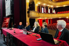 Na scenie zabytkowego teatru przy długim stole z bordowym obrusem siedzi pięć osób z laptopami i mikrofonami, zwrócone w stronę widowni. W tle rzędy czerwonych foteli i złocone balkony, po lewej na sztaludze plakat festiwalowy.