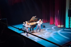 Na scenie występuje trzech muzyków: dwóch skrzypków stoi na przodzie i gra z energią, a za nimi pianista siedzi przy dużym, otwartym fortepianie. Artyści są oświetleni delikatnymi reflektorami, które tworzą jasne wzory na podłodze.