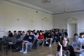 Sala szkoleniowa z grupą uczniów uczestniczących w kursie w ramach projektu unijnego. Młodzież siedzi na krzesłach, trzymając materiały edukacyjne. W tle widoczne drzwi, białe ściany i tablica informacyjna o dofinansowaniu UE.
