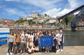 Grupa młodzieży i kilka dorosłych osób pozujących nad rzeką w Portugalii. W tle widać kolorowe kamienice i most. Jedna z dziewczyn trzyma flagę Unii Europejskiej. Zdjęcie zrobiono w słoneczny dzień.