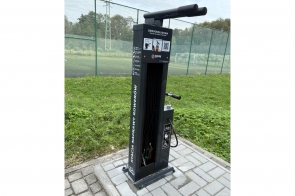 Czarna samoobsługowa stacja naprawy rowerów z wiszącymi narzędziami i pompką, ustawiona na kostce brukowej przy ogrodzonym boisku, wokół trawa.