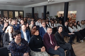 Sala wypełniona siedzącymi ludźmi, głównie dorosłymi i młodzieżową, ustawionymi w rzędach. Wszyscy są elegancko ubrani i skierowani w jedną stronę. W tle znajdują się regały z książkamioraz kolumny.