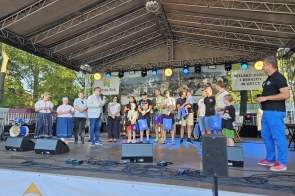 Grupa osób stoi na scenie plenerowej – część w strojach regionalnych, część w sportowych ubraniach lub casualu. Trzymają torby z upominkami. W tle baner z napisem „Bielsko-Biała i Beskidy w Ustce”.