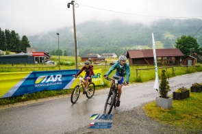 Dwójka rowerzystów na rowerach górskich, jadących po mokrej szosie.W tle zamglony i zalesiony stok. 