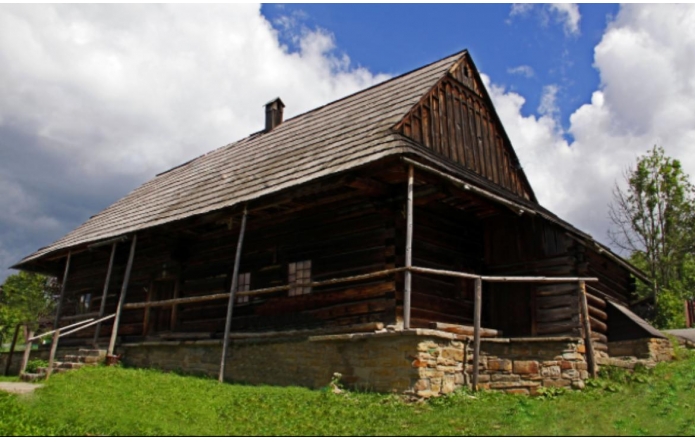 Drewniana, zabytkowa chata góralska na kamiennej podmurówce, z szerokim gankiem i balustradą z surowych żerdzi. Spadzisty, gontowy dach i ciemne, ciosane bale podkreślają tradycyjny beskidzki charakter. Wokół zielona trawa, w tle niebo z kłębami chmur.