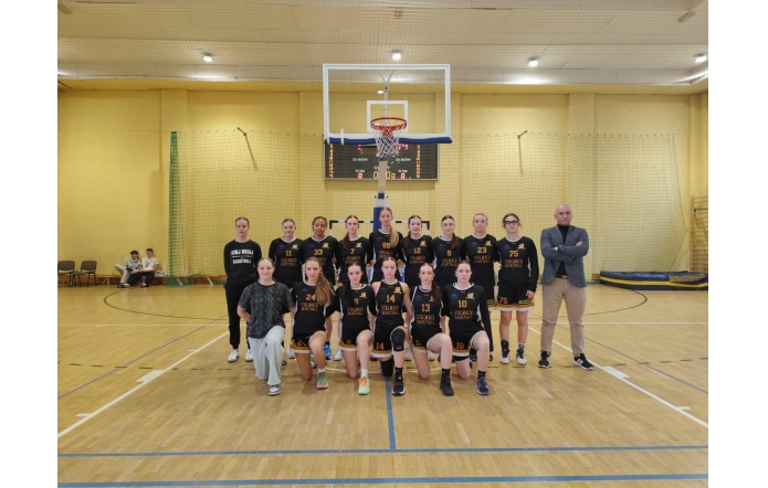 Drużyna koszykarek Liceum Ogólnokształcącego im. Pawła Stalmacha w Wiśle wraz z trenerami pozuje na hali sportowej pod koszem. Zawodniczki w czarnych strojach z napisem „Stalmach Basketball” prezentują się w sportowej, drużynowej atmosferze.