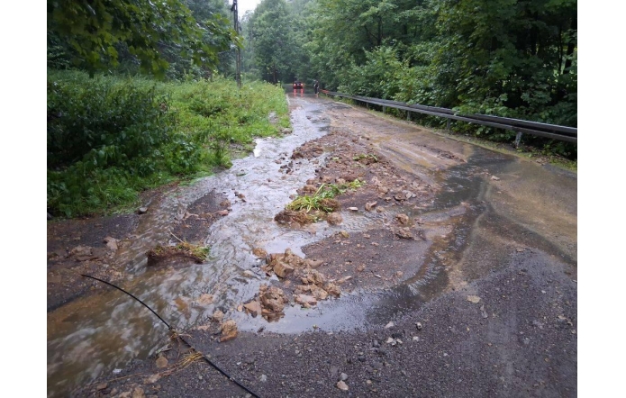 Rozmyta nawierzchnia drogi z zalegającym rumoszem i wypływającą wodą opadową. Widoczne zniszczenia i zalanie pobocza w wyniku intensywnych opadów w zalesionym terenie. W tle ustawione barierki ostrzegawcze.