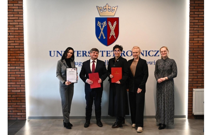 Sześć osób stojących w rzędzie w holu budynku, na tle jasnej ściany z herbem. Trzy osoby trzymają czerwone teczki, jedna osoba trzyma certyfikat w białej oprawie. Uczestnicy ubrani są w stroje formalne. Po bokach ściany widoczne fragmenty ceglanej ściany oraz grzejnik.