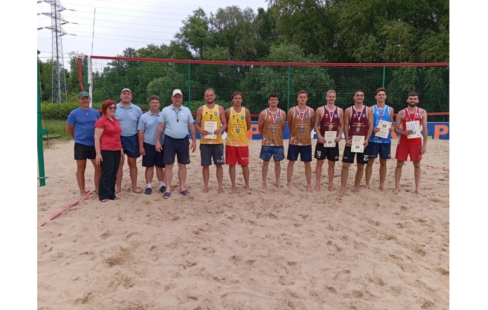 iGrupa osób stoi na boisku do siatkówki plażowej. Pośrodku uśmiechnięci zawodnicy w sportowych strojach prezentują medale i dyplomy. Obok nich organizatorzy w koszulkach polo. W tle siatka i drzewa, atmosfera sportowego święta.