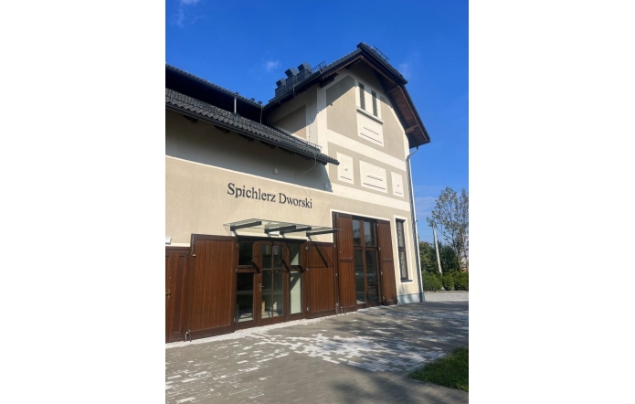 Budynek o jasnej elewacji z ciemnym dachem, dużymi drewnianymi drzwiami i przeszklonymi oknami na parterze. Przed budynkiem znajduje się kamienny podjazd, a w tle widać drzewa i niebo.