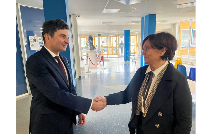 Dwójka osób w oficjalnych strojach podaje sobie dłonie w przestronnym, dobrze oświetlonym holu. Marek Wójcik Wojewoda Śląski  po lewej ma na sobie ciemny garnitur, białą koszulę i bordowy krawat. Janina Żagan Starosta Cieszyński po prawej ubrana jest w elegancką czarną marynarkę z ozdobnymi lamówkami oraz białą koszulę.