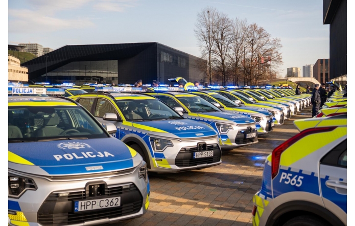 Rząd nowych policyjnych radiowozów stoi ustawiony w równych liniach na otwartej przestrzeni w słoneczny dzień. Samochody mają biało-niebieskie malowanie z żółtymi elementami odblaskowymi oraz włączone sygnały świetlne. W tle widać nowoczesne budynki, funkcjonariuszy oraz bezlistne drzewa.