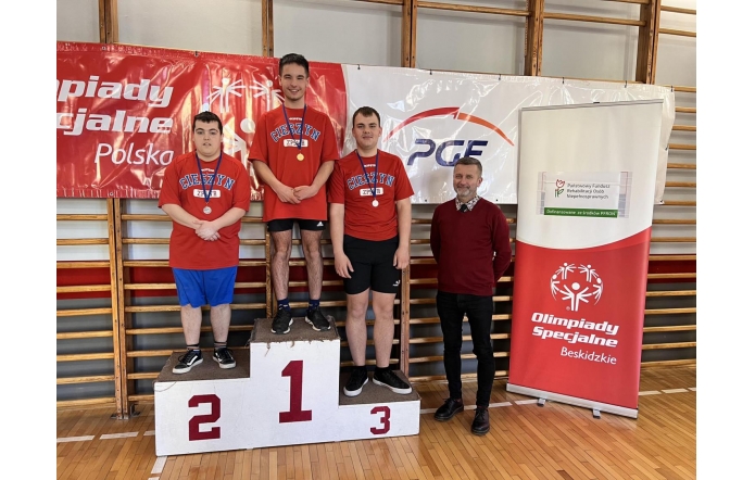 Trzech młodych sportowców z medalami na podium po zawodach Olimpiad Specjalnych. W tle banery z logo wydarzenia, obok podium stoi uśmiechnięty mężczyzna w bordowym swetrze (Michał Rajwa, Naczelnik Wydziału Edukacji Starostwa Powiatowego w Cieszynie). Wszyscy ubrani w czerwone koszulki z napisem „Cieszyn ZPSWR”.