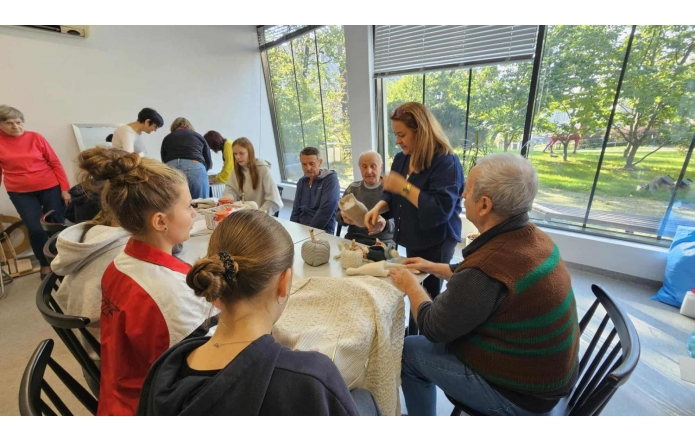 Wokół stołu siedzi osiem osób - trzech mężczyzn i piątka dziewcząt,a jedna kobieta stoi przy stole, na którym leżą elementy do tworzenia rękodzieła.W pomieszczeniu stoją jeszcze cztery kobiety.Jedna ze scian jest całkowicie przeszklona,za nią widać trawnik i drzewa.