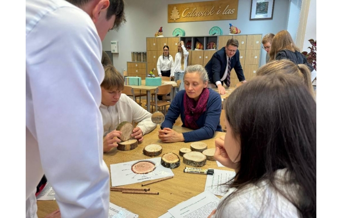Przy stole siedzi grupa uczniów, którzy trzymają w dłoniach drewniane krążki i oglądają je z bliska. Na blacie leży kilka kolejnych fragmentów drewna, kartki z zadaniami oraz przybory do pisania. Obok nich siedzi dorosła kobieta, patrząca w kierunku krążków. W tle widać inne osoby pracujące w sali oraz szafki