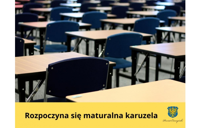 Sala egzaminacyjna wypełniona pustymi, równomiernie ustawionymi ławkami i granatowymi krzesłami, przygotowana na rozpoczęcie egzaminów maturalnych. Na dole żółty pasek z napisem „Rozpoczyna się maturalna karuzela” oraz herbem Powiatu Cieszyńskiego.