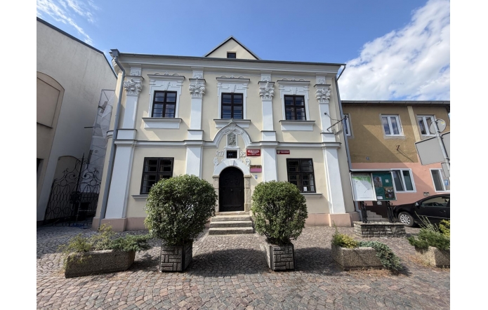 Zabytkowy, jasny budynek z ozdobną fasadą i ciemnymi oknami. Przed wejściem znajdują się schody, krzewy w donicach oraz brukowany plac, a w tle widać błękitne niebo.