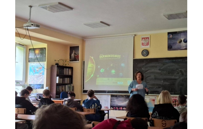 Uczniowie siedzą w sali lekcyjnej, słuchając prelegentki prowadzącej zajęcia w ramach programu „Dermokosmos”. Na ekranie widoczna prezentacja z planetami i logotypami organizatorów. W tle tablica i godło Polski.