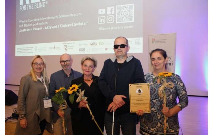 Pięć osób stoi na tle ekranu z logo konferencji REHA FOR THE BLIND IN POLAND. Troje z nich trzyma słoneczniki, jedna osoba trzyma dyplom. Wszyscy są uśmiechnięci, ubrani elegancko. W tle wyświetlone są logotypy partnerów wydarzenia.