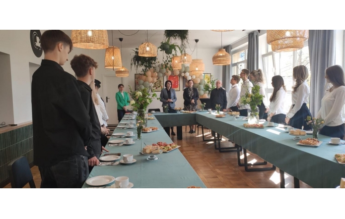 Uczniowie i nauczyciele stoją przy elegancko nakrytym, ułożonym w kształt litery U stole, udekorowanym kwiatami i przekąskami. Wnętrze o ciepłym, naturalnym wystroju z wiklinowymi lampami i pastelowymi balonami sprzyja uroczystości.