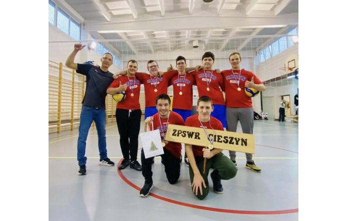Grupa zawodników w czerwonych koszulkach sportowych pozuje w sali gimnastycznej z medalami na szyjach, uśmiechając się do obiektywu, trzymając piłki siatkarskie oraz tabliczkę z nazwą ZPSWR Cieszyn.