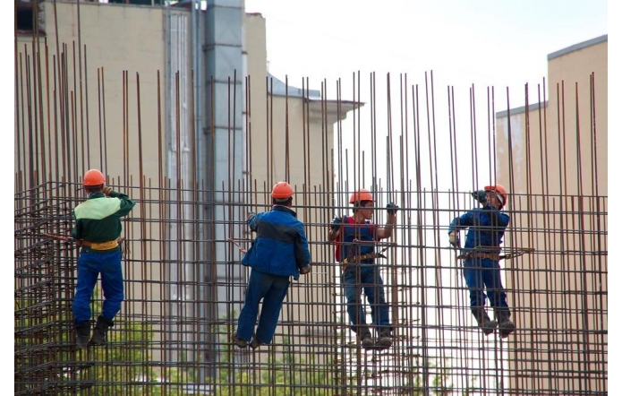 Czterech pracowników ubranach w robocze kombinezony i pomarańczowe kaski wykonuje prace na wysokości, wspinając się po metalowej konstrukcji złożonej z gęsto rozmieszczonych stalowych prętów. Każdy z nich jest przypięty uprzężą asekuracyjną. W tle widać wysokie budynki, które sugerują, że prace odbywają się w miejskim otoczeniu. Ich precyzyjne ruchy świadczą o doświadczeniu i koncentracji.