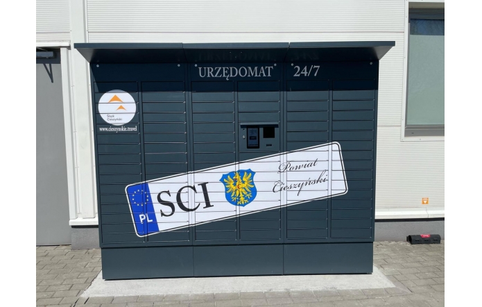 Urzędomat zlokalizowany przy budynku urzędu, oznaczony napisem „URZĘDOMAT 24/7”. Urządzenie ma ciemny kolor, z grafiką przypominającą tablicę rejestracyjną z napisem „SCI” i herbem Powiatu Cieszyńskiego.