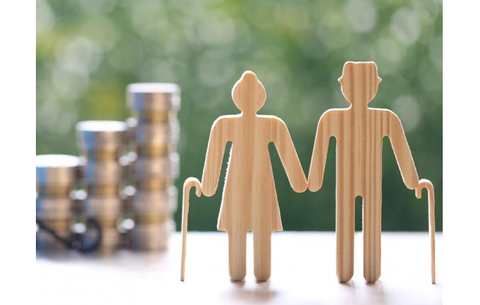 Dwie drewniane sylwetki starszych osób trzymają się za ręce i opierają na laskach, a w tle widać stosy monet oraz rozmytą zieleń.