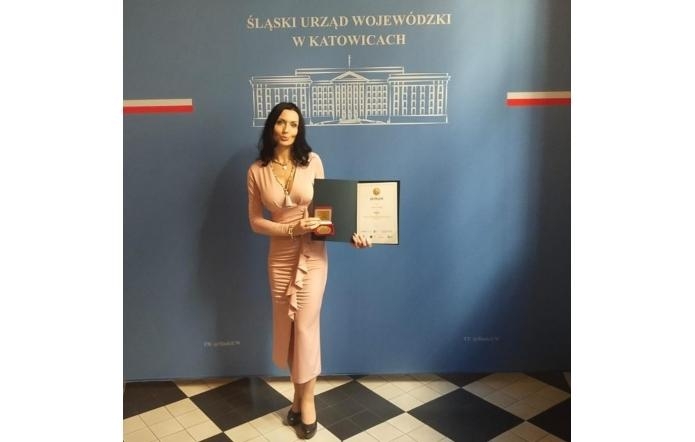 Agata Gacon w eleganckiej, różowej sukni trzyma dyplom i medal na tle ścianki z napisem „Śląski Urząd Wojewódzki w Katowicach”. Uhonorowana w oficjalnym wnętrzu, uśmiecha się z dumą, prezentując swoje wyróżnienie.
