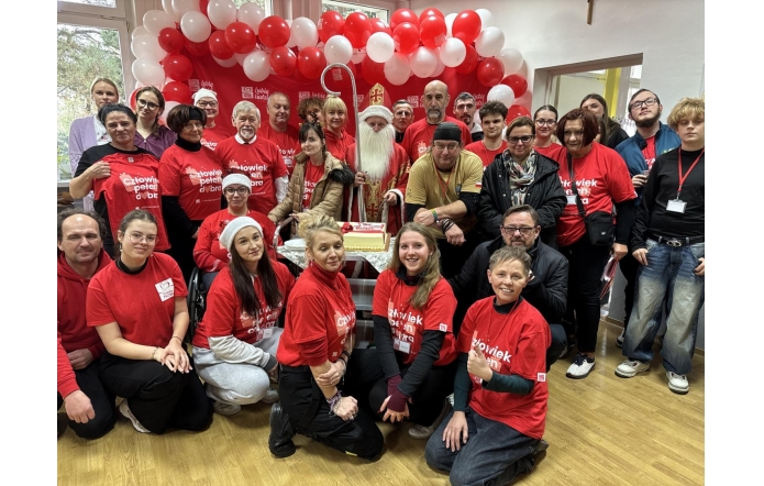 Grupa kilkudziesięciu osób w czerwonych koszulkach z białymi napisami, pozujących wspólnie w sali ozdobionej czerwono-białymi balonami, z osobą ubraną w strój św. Mikołaja stojącą pośrodku, przy stole z tortem.