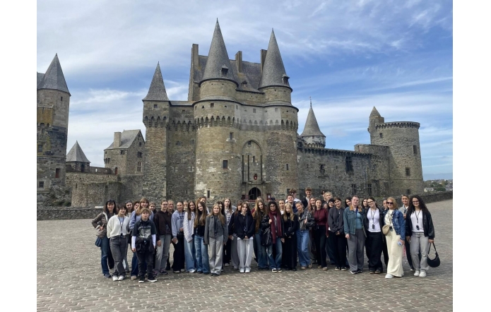 Grupa młodych osób stoi razem przed dużym średniowiecznym zamkiem z kamienia. Budowla ma kilka wysokich, stożkowatych wież i grube mury obronne z wąskimi oknami. Niebo jest jasne, z lekkimi chmurami, a plac, na którym stoją, jest wyłożony brukiem. Ludzie uśmiechają się i pozują do wspólnego zdjęcia.