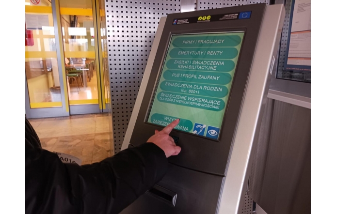 Widoczna ręka osoby w ciemnej kurtce dotyka palcem ekranu dotykowego automatu do obsługi klientów w urzędzie. Na ekranie wyświetlone jest menu wyboru spraw administracyjnych, w tym emerytury, zasiłki, świadczenia rodzinne oraz opcja „wizyta zarezerwowana”. W tle przeszklone drzwi i fragment poczekalni.