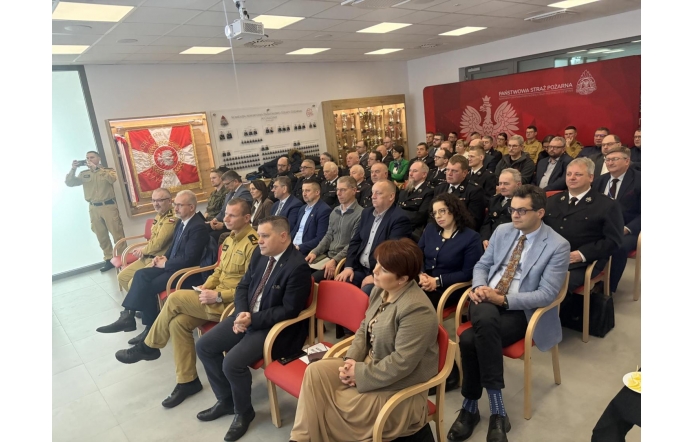 Sala konferancyjna z rzędami krzeseł, na których siedzi kilkadziesiąt osób, część w mundurach strażackich, a część w eleganckich strojach. W tle czerwona ścianka z godłem i napisem " Państwowa Straż Pożarna", obok przeszklona gablota z pucharami. Na suficie projektor.