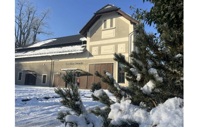Jasny budynek z dwuspadowym dachem, częsciowo pokrytym śniegiem, widoczny na tle niebieskiego nieba. Przed budynkiem leży warstwa śniegu, a na pierwszym planie znajdują się gałęzie sosny z osadzonym śniegiem. Na elewacji widczony napis "Spichlerz Dworski"