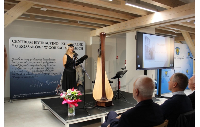 Dyrektor CEK "U KOSSAKÓW" Urszula Broda Gawełek w czarnej sukni, długich rękawiczkach i ozdobnej opasce z piórami przemawia do mikrofonu na scenie. Przed nią stoi harfa i wazon z bukietem kwiatów owiniętym różową wstążką. W tle widoczna jest tablica z napisem „Centrum Edukacyjno-Kulturalne U Kossaków w Górkach Wielkich” oraz ekran z prezentacją zatytułowaną „Kossakowie – życie w cieniu ruin”. Publiczność siedzi naprzeciw, uważnie słuchając wystąpienia.