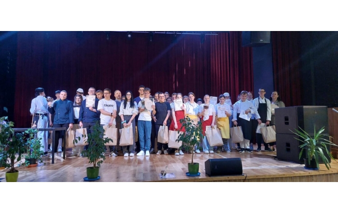 Grupa osób stoi na scenie, część z nich trzyma dyplomy, statuetki oraz papierowe torby. W tle widoczna jest bordowa kurtyna, a po bokach sceny ustawiono głośniki. Przed sceną znajdują się rośliny doniczkowe oraz elementy wyposażenia technicznego.